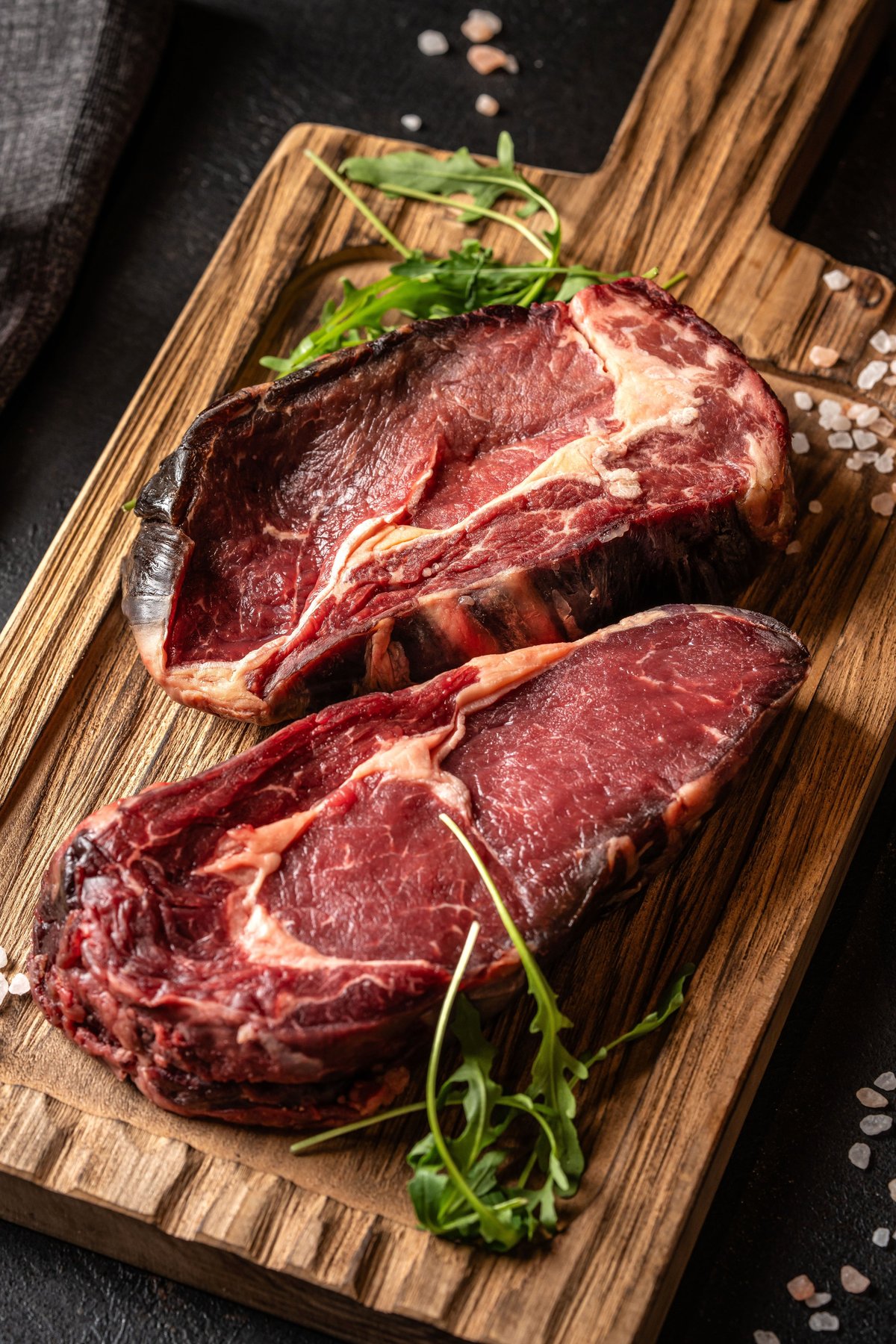 Close-up of raw dry aged ribeye steak on rustic wooden cutting board with pink salt and arugula leaves.
